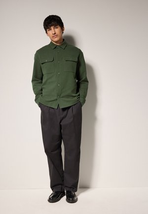 Homme debout contre un mur beige portant une chemise verte boutonnée, un pantalon ample sombre et des mocassins noirs, mains dans les poches.