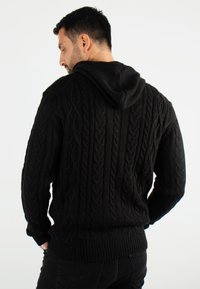 Hoodie en maille noire avec un motif torsadé texturé, des poignets et un ourlet côtelés. Le tissu présente une apparence douce, mettant en valeur un design décontracté.
