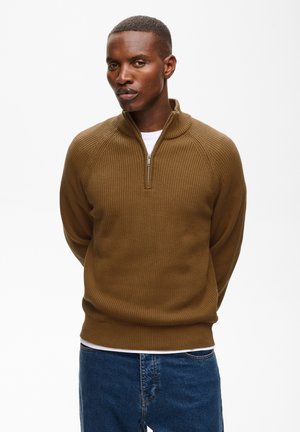 Jeune homme aux cheveux courts portant un pull en maille marron zippé et un jean bleu, debout les mains dans le dos sur fond blanc.