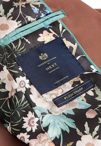 Tissu à motif floral avec bordure bleu sarcelle, étiquettes bleu marine indiquant "NEXT Skinny Fit Made in China" et "#LOOKSHARP" sur un fond marron.