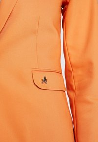 Gros plan sur la manche et la poche d'un blazer orange avec une petite épingle métallique décorative "M" sur le rabat de poche.
