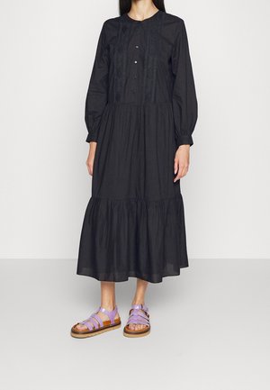 Frau trägt langes schwarzes Kleid mit Knöpfen und Spitzendetails sowie lila flachen Sandalen, stehend vor schlicht weißem Hintergrund.