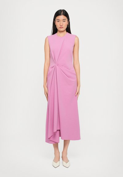 Robe sans manches rose avec un design tordu à la taille, une silhouette fluide et une longueur en dessous du genou. Portée avec des chaussures blanches à bout pointu.