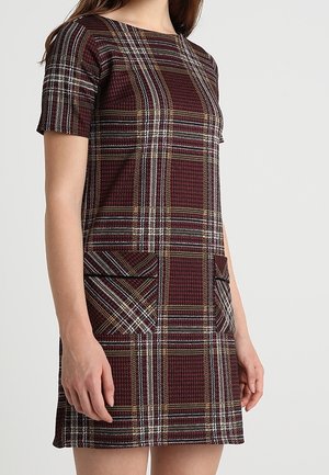 Femme portant une robe à manches courtes, longueur aux genoux, avec un motif à carreaux bordeaux, marron et blanc, et deux poches avant à rayures diagonales.
