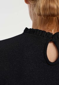 Rückenansicht einer Person mit blonden Haaren, die ein schwarzes, strukturiertes Oberteil mit einem Rüschenausschnitt und einem Schlüssellochausschnitt im Nacken trägt.
