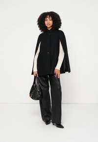 Μαύρο παλτό-κάπα με στρογγυλό γιακά, κλείσιμο στο μπροστινό μέρος με κουμπιά, πάνω από λευκό πλεκτό πουλόβερ, συνδυασμένο με γυαλιστερό μαύρο δερμάτινο παντελόνι και μαύρη τσάντα χειρός.