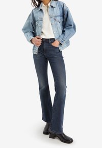Chaqueta de mezclilla azul clara sobre una polo blanca, combinada con jeans acampanados azul oscuro y botas de tacón negras. Texturas suaves, diseño casual.
