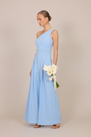 Γυναίκα με ανοιχτό μπλε ολόσωμη φόρμα με έναν ώμο, κρατώντας λευκές κάλλες με κορδέλα, φορώντας χρυσά σκουλαρίκια και σανδάλια.