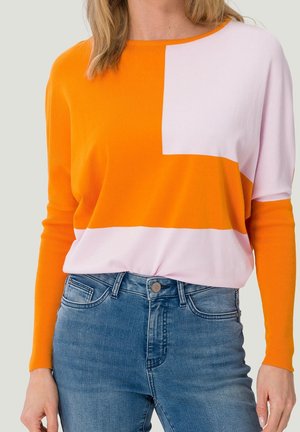 Femme portant un pull à manches longues à blocs de couleurs orange et rose clair avec un jean bleu, sur un fond neutre.