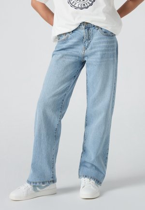 Personne portant un jean droit bleu clair, des baskets blanches et un t-shirt blanc partiellement visible avec un motif imprimé.