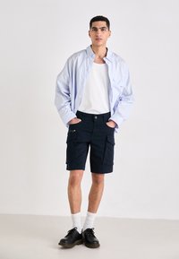 Chemise boutonnée à rayures bleu clair sur un débardeur blanc, associée à un short cargo noir et des chaussures noires. Le short a plusieurs poches.