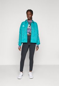 Kvinna står klädd i en turkosmönstrad jacka, en färgglad topp med fjärilstryck, svarta leggings och vita sneakers mot en enkel ljus bakgrund.