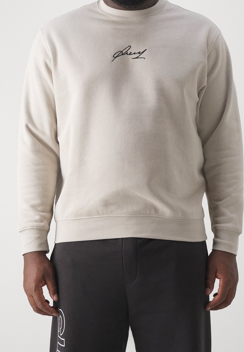 Felpa beige chiaro con maniche lunghe, scollo rotondo e firma ricamata nera sul petto; abbinata a pantaloni grigio scuro.