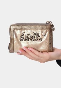 Monedero de cuero sintético dorado con acabado texturizado, que presenta un llamativo logo "AneKKe" y un acento en forma de corazón. Cierre con cremallera y correa para la muñeca.