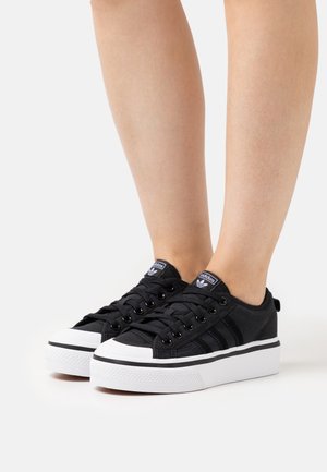 Nogi w czarnych niskich butach sportowych Adidas z białymi grubymi podeszwami i czarnymi sznurowadłami, stojące na białej powierzchni.