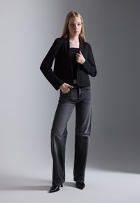 Černý zkrácený blazer přes černý top, doplněný šedými širokými džínami a ostrými černými podpatky, na pozadí s gradientem šedé barvy.