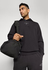 Black fabric hooded sweatshirt with drawstrings and logo, paired with matching sweatpants. Model holds a black zippered bag.
