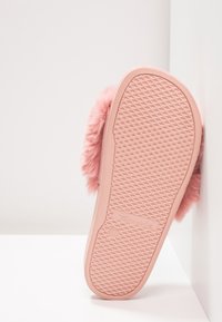 Pantoufle rose unique avec tige duveteuse, vue par la semelle, appuyée contre un mur et une surface blancs.