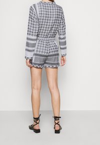 Šedý vzorovaný overal s dlouhými rukávy, elastickým pasem a šortkami. Obsahuje geometrické vzory, různé textury a kontrastní linie. Černé sandály.