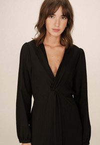 Robe noire en tissu léger, dotée d'un décolleté en V profond, de longues manches bouffantes et d'un détail de nœud froncé à la taille.