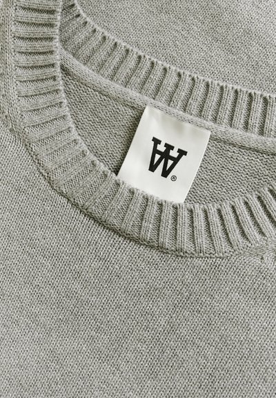 Grå strikket sweater med ribbet rund hals og blød tekstur, med et label der viser brandets logo i sort på en hvid baggrund.