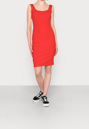 Kobieta w czerwonej sukience bez rękawów sięgającej kolan oraz czarno-białych trampkach, stojąca na jasnym, jednolitym tle.