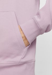 Sudadera de color púrpura claro con bolsillo tipo canguro, puños acanalados y textura suave. Vista en primer plano que resalta las costuras y detalles de la tela.
