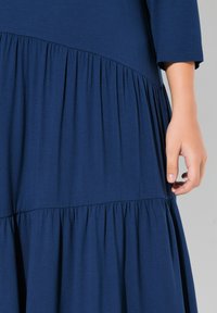 Abito blu navy con un design a strati, caratterizzato da una parte superiore liscia e un fondo arricciato. Tessuto morbido con una lucentezza delicata. Maniche corte, mano mostrata.