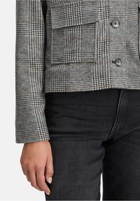 Zbliżenie na osobę w szarym, w kratkę żakiecie z guzikami i kieszenią klapkową, w połączeniu z czarnymi dżinsami, z ręką swobodnie opartą na boku.