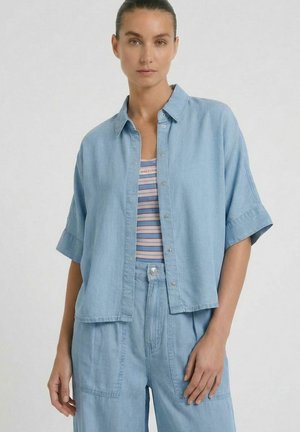 Vrouw draagt een losse lichtblauwe korte-mouwen blouse met knopen over een gestreepte tanktop en bijpassende lichtblauwe hooggetailleerde broek.