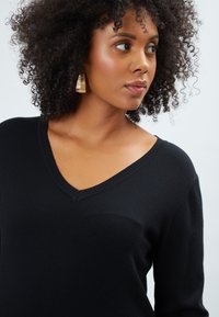 Maglione nero a V con un motivo a maglia testurizzato, dotato di maniche lunghe e una vestibilità rilassata. Il modello indossa orecchini a cerchio dorati.