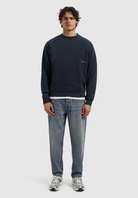 Navyblauwe sweatshirt met ronde hals, zachte textuur en klein logo. Gecombineerd met lichtblauwe rechte jeans en grijze sneakers.