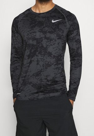 Homme portant un t-shirt de compression Nike Pro à manches longues noir et gris à motifs, avec un short noir, mains dans les poches, debout devant un fond uni.