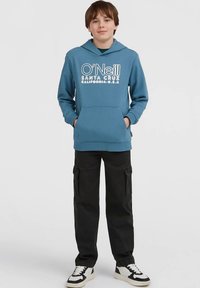 Sudadera con capucha de algodón azul con texto gráfico blanco "O'Neill Santa Cruz" en el pecho, bolsillo frontal, capucha ajustable, combinada con pantalones cargo negros y zapatillas.