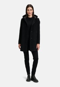 Zwarte jas met een gestructureerde afwerking, capuchon en twee knopen. Draagt over een zwarte outfit met slim-fit broek en enkellaarsjes.