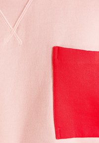 Tela de sudadera rosa con un bolsillo parche texturizado en color rojo brillante. Presenta detalles de costura en V y una textura suave y lisa.