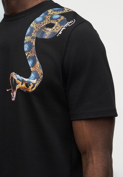 Μαύρο μπλουζάκι με γραφικό ένας φιδιού σε μπλε, κίτρινο και πορτοκαλί που τυλίγεται από το στήθος στον ώμο, φορεμένο από άτομο με σκούρο δέρμα.