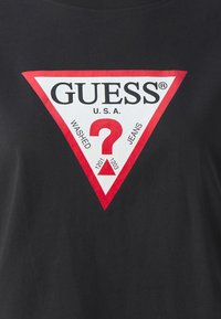 Juodas marškinėliai su dideliu raudonu ir baltu trikampiu logotipu. Tekstas apima "GUESS", "U.S.A.", "WASHED" ir "JEANS" su klausimo ženklu.
