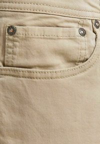 Il tessuto di cotone beige presenta una tasca posteriore con cuciture a contrasto e rivetti in metallo, mostrando una texture liscia e un design semplice.