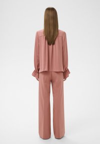 Kvinde med langt glat hår iført en støvet lyserød bluse med lange ærmer og brede bukser, stående ryggen til på en hvid baggrund.