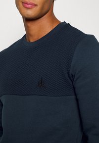 Sudadera azul marino con panel de punto texturizado en la parte superior, tela lisa en la parte inferior y un pequeño logo negro en el pecho.