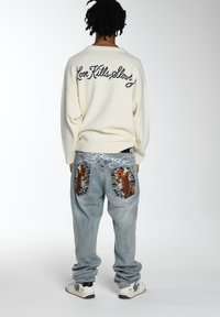Weißer Pullover mit "Love Kills Slowly" in schwarz gestickt, kombiniert mit hellblauen Jeans aus Denim, die mit gestickten orangen Koi-Fisch-Patches verziert sind.