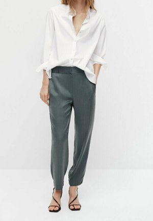 Femme portant une chemise blanche ample à boutons, un pantalon gris fuselé et des sandales à talons noires à brides, debout devant un fond blanc uni.