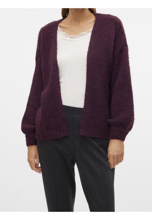 Femme portant un cardigan en maille violet ample et ouvert sur le devant, par-dessus un haut blanc bordé de dentelle et un pantalon gris foncé, vue des épaules aux hanches.