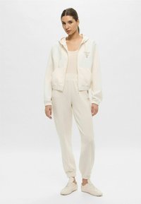 Felpa con zip color beige chiaro, dotata di tasche frontali e cappuccio con cordino, abbinata a pantaloni della tuta tapered coordinati e sneakers color crema.