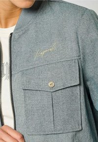 Light gray denim jacket with a zippered front, featuring a chest pocket with a metal button and cursive logo embroidery. Textured fabric.