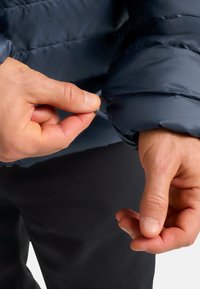 Veste matelassée navy avec un design texturé et en relief. Une main ajuste la manche, révélant des poignets élastiques et un détail à cordon de serrage.