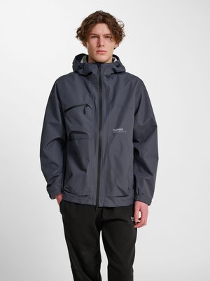 Jeune homme portant une veste imperméable gris foncé avec capuche et pantalon noir, debout devant un fond clair uni.