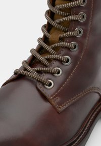 Botín de cuero marrón con puntera redonda, presenta cordones texturizados en marrón y blanco, ojales de metal y detalles de costura en contraste.
