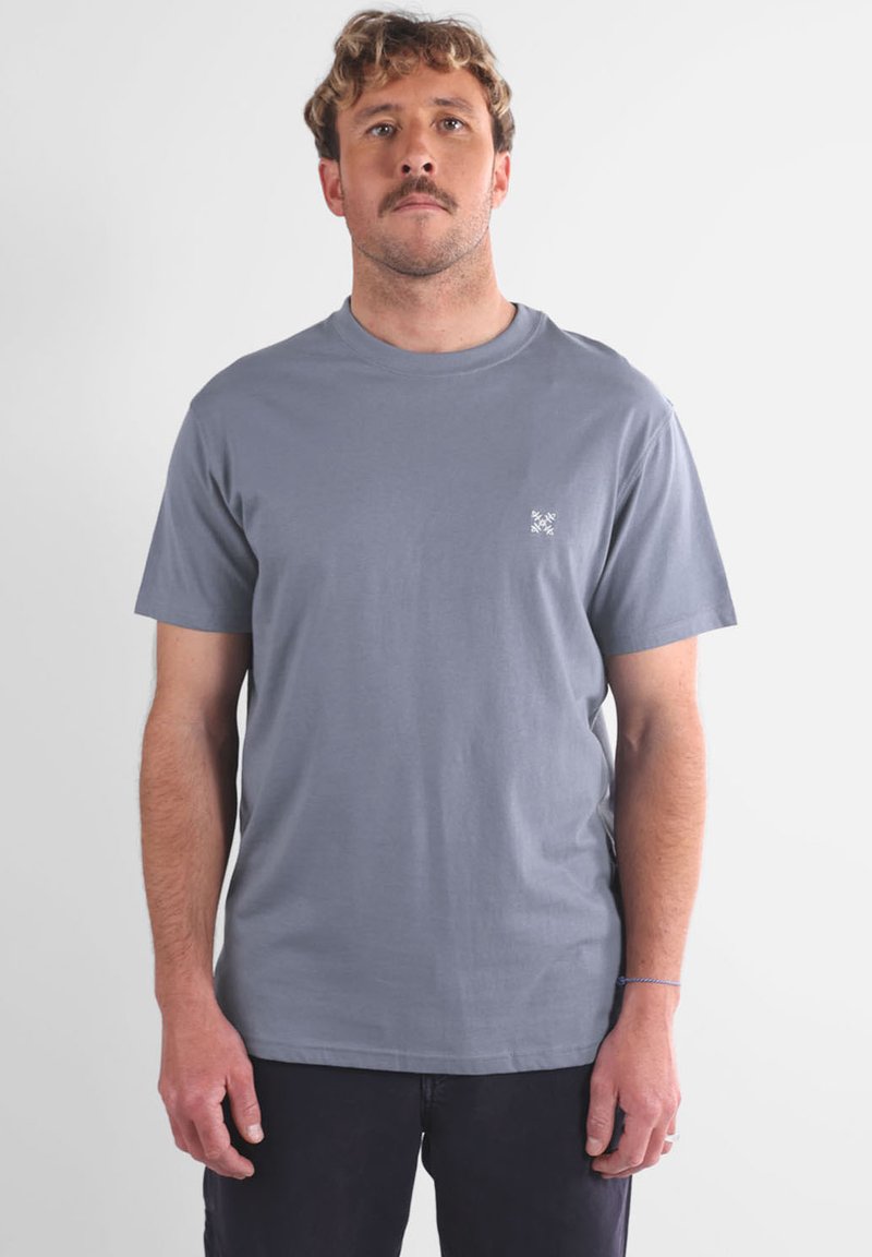 Homme aux cheveux châtains clairs et à la moustache, portant un t-shirt gris unis à manches courtes et un pantalon sombre, se tenant contre un fond blanc.